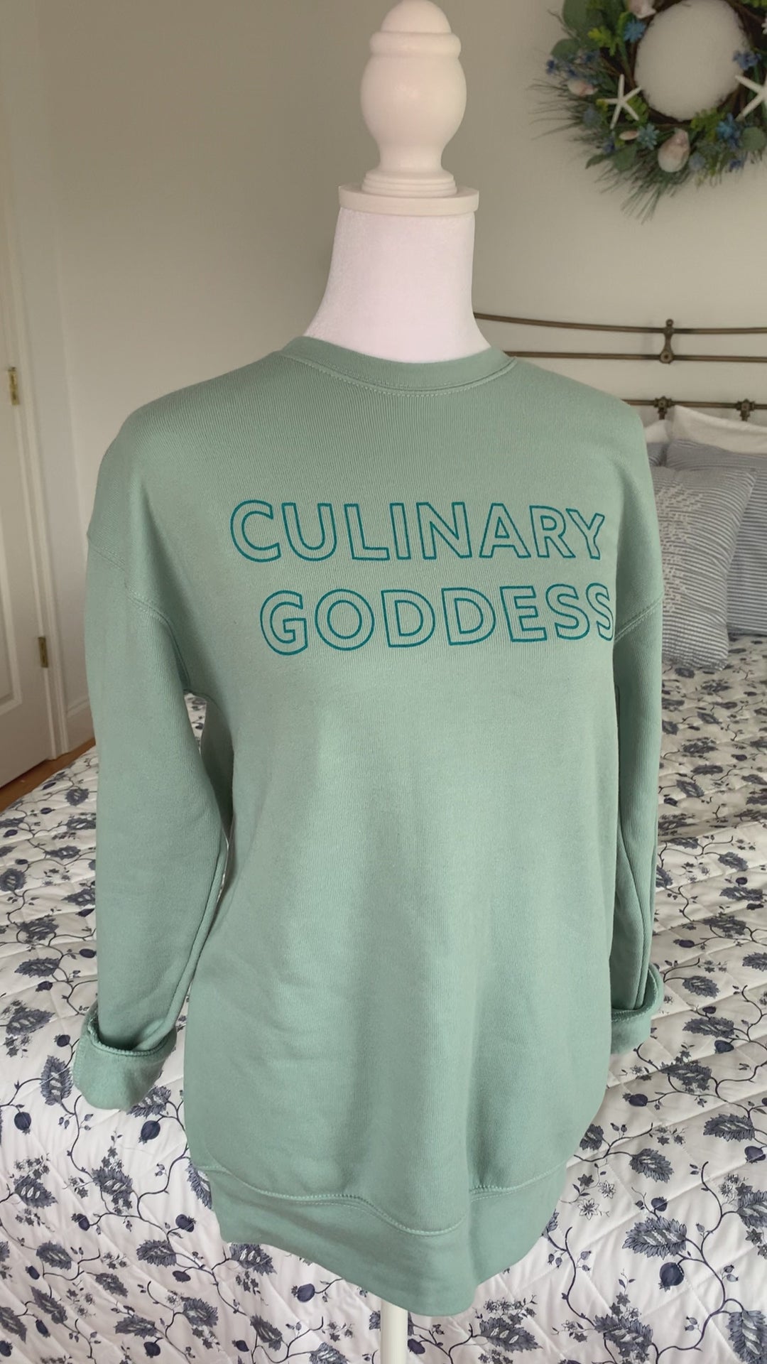 A dusty blue crewneck sweatshirt with the words "Culinary Goddess" hangs on a manikin