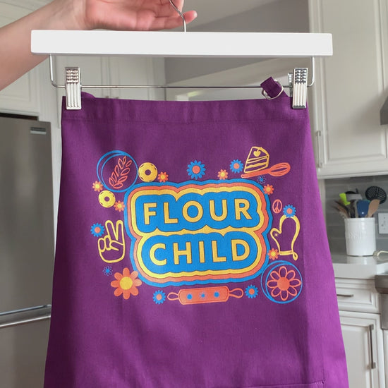 A purple apron that reads "Flour Child" with colorful illustrations hangs on a hanger