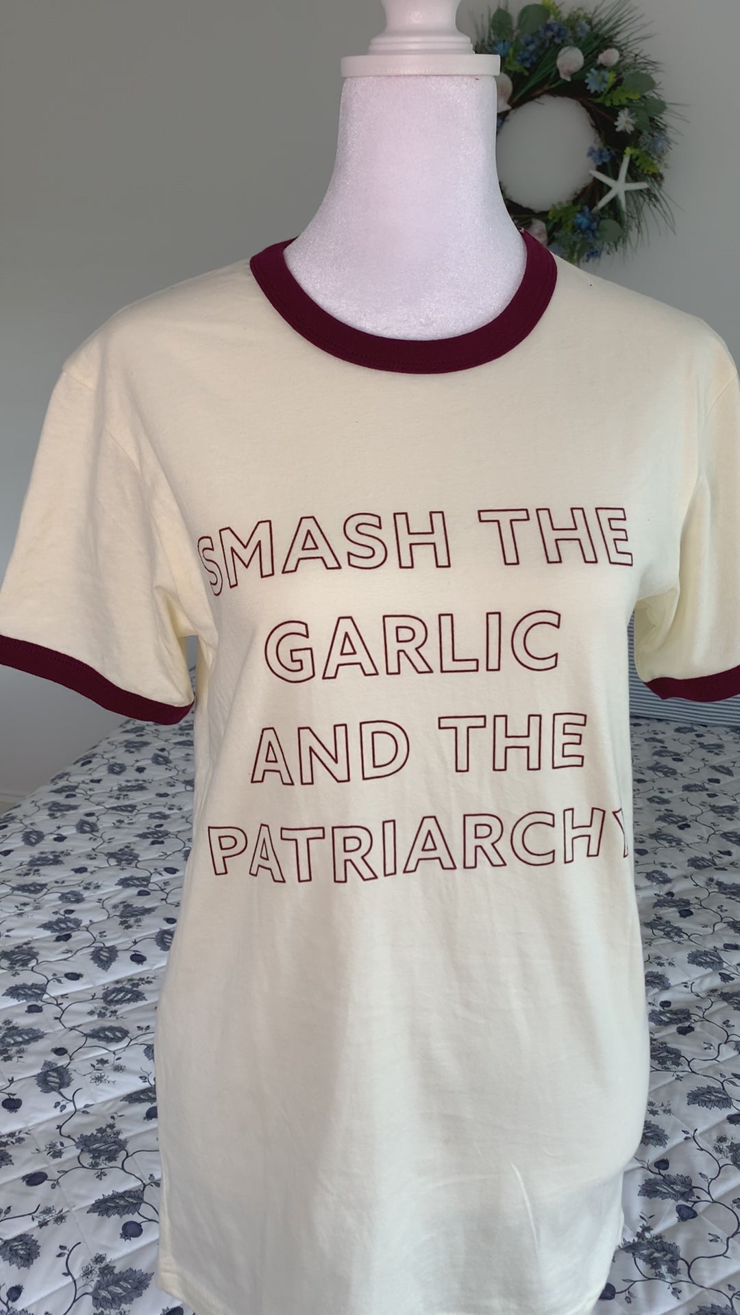 An off-white ringer tee that reads "Smash the Garlic and the Patriarchy" hangs on a manikin