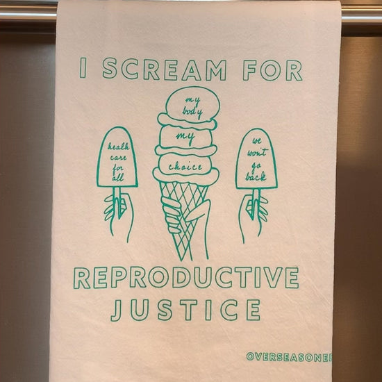 A white tea towel with mint green block letters that reads "I scream for reproductive justice" hangs in a kitchen
