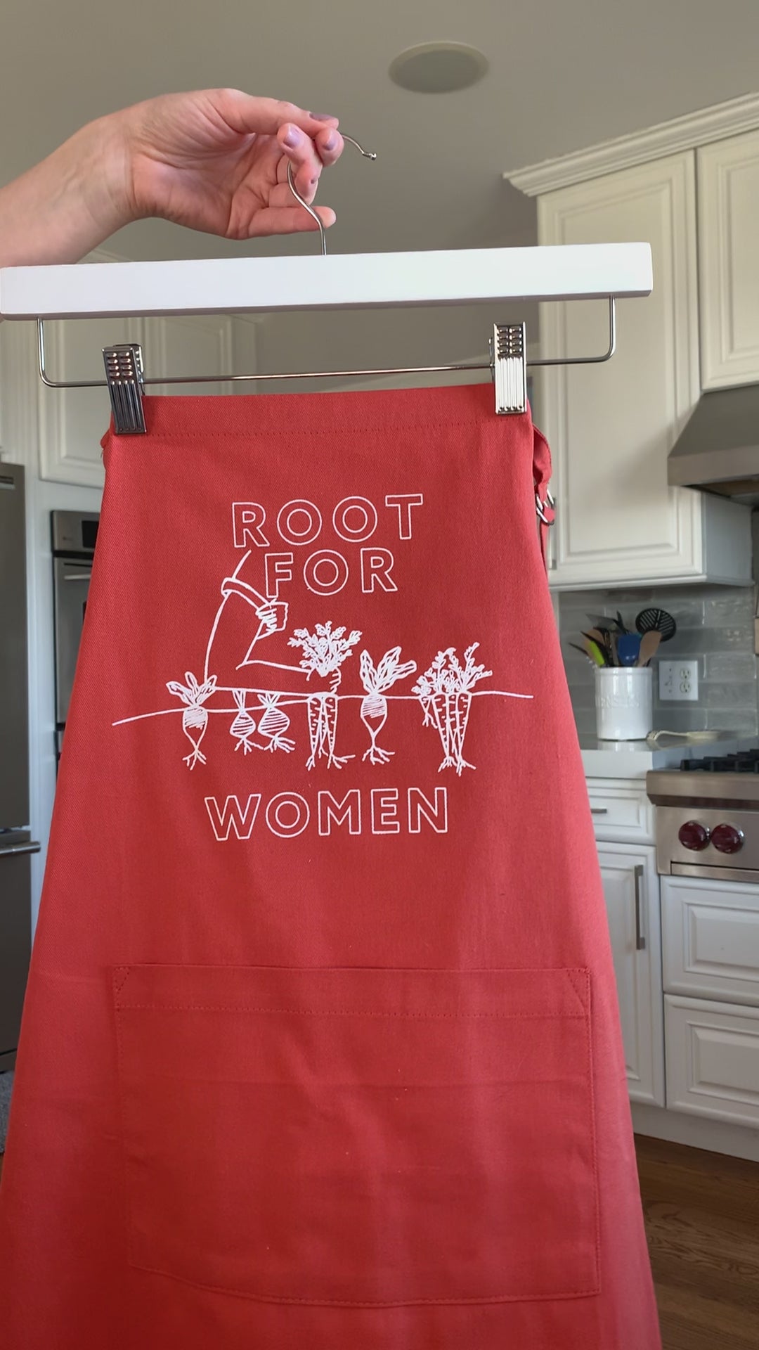 An orange apron that reads "Root for Women" in white block letters hangs on a hanger