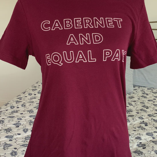A maroon t-shirt that reads "Cabernet and Equal Pay" hangs on a manikin
