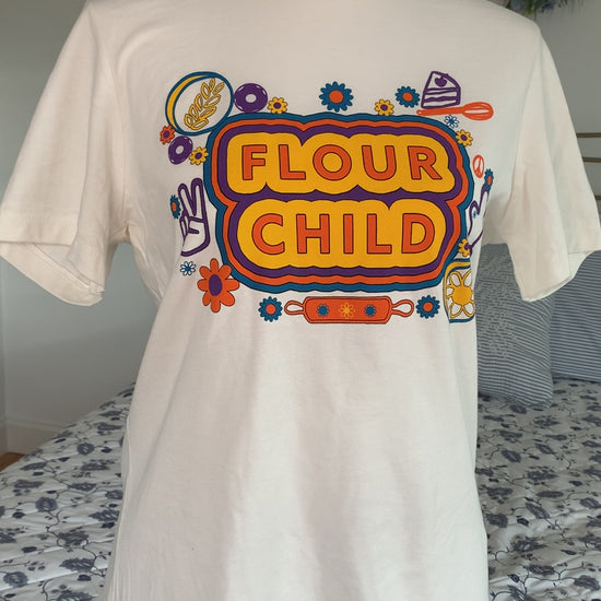 A white t-shirt that reads "Flour Child" with colorful designs hangs on a manikin