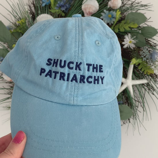 A woman holds a light blue baseball hat that reads "Shuck the Patriarchy"