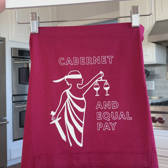 A red apron that reads "Cabernet and Equal Pay" hangs on a hanger