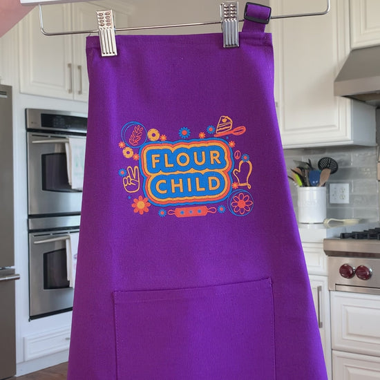 A purple kid's apron that reads "Flour Child" hangs on a hanger