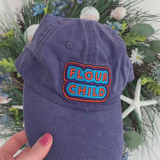 A woman holds a blue baseball cap with embroidery that reads "Flour Child"
