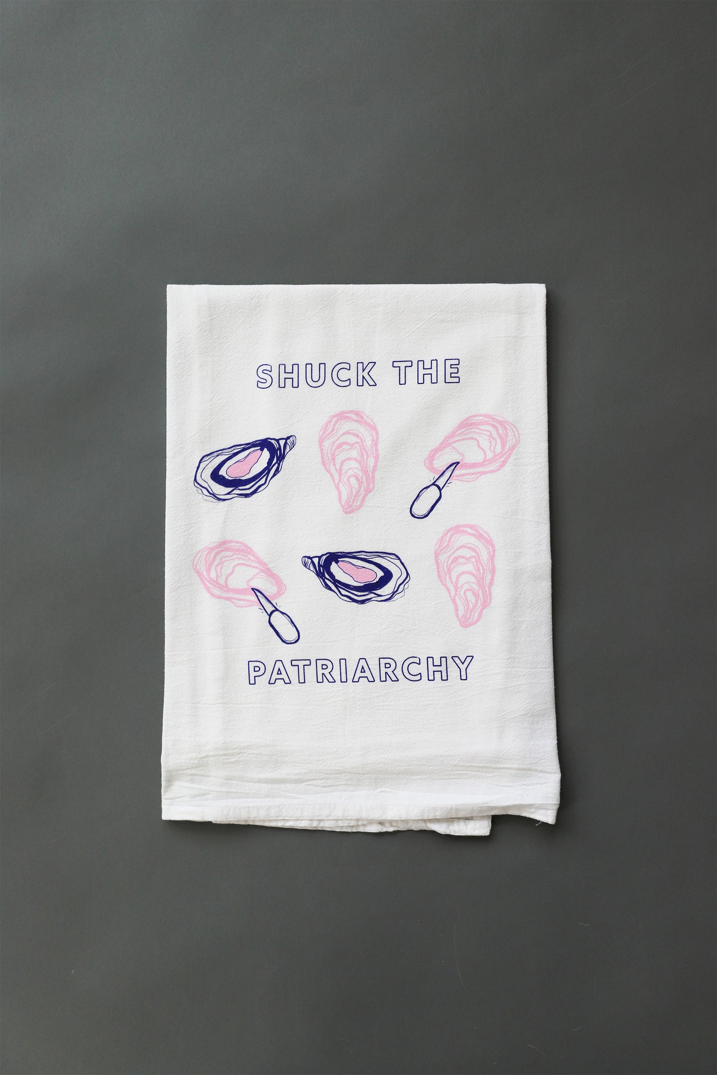 A white tea towel with "Shuck the Patriarchy" in blue block letters and pink and blue oyster illustrations