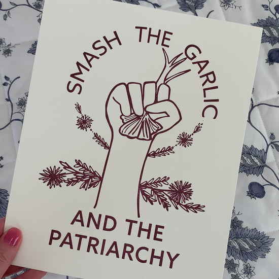 An art print that reads "Smash the Garlic and the Patriarchy" in a dark red color with an illustration of a hand holding garlic