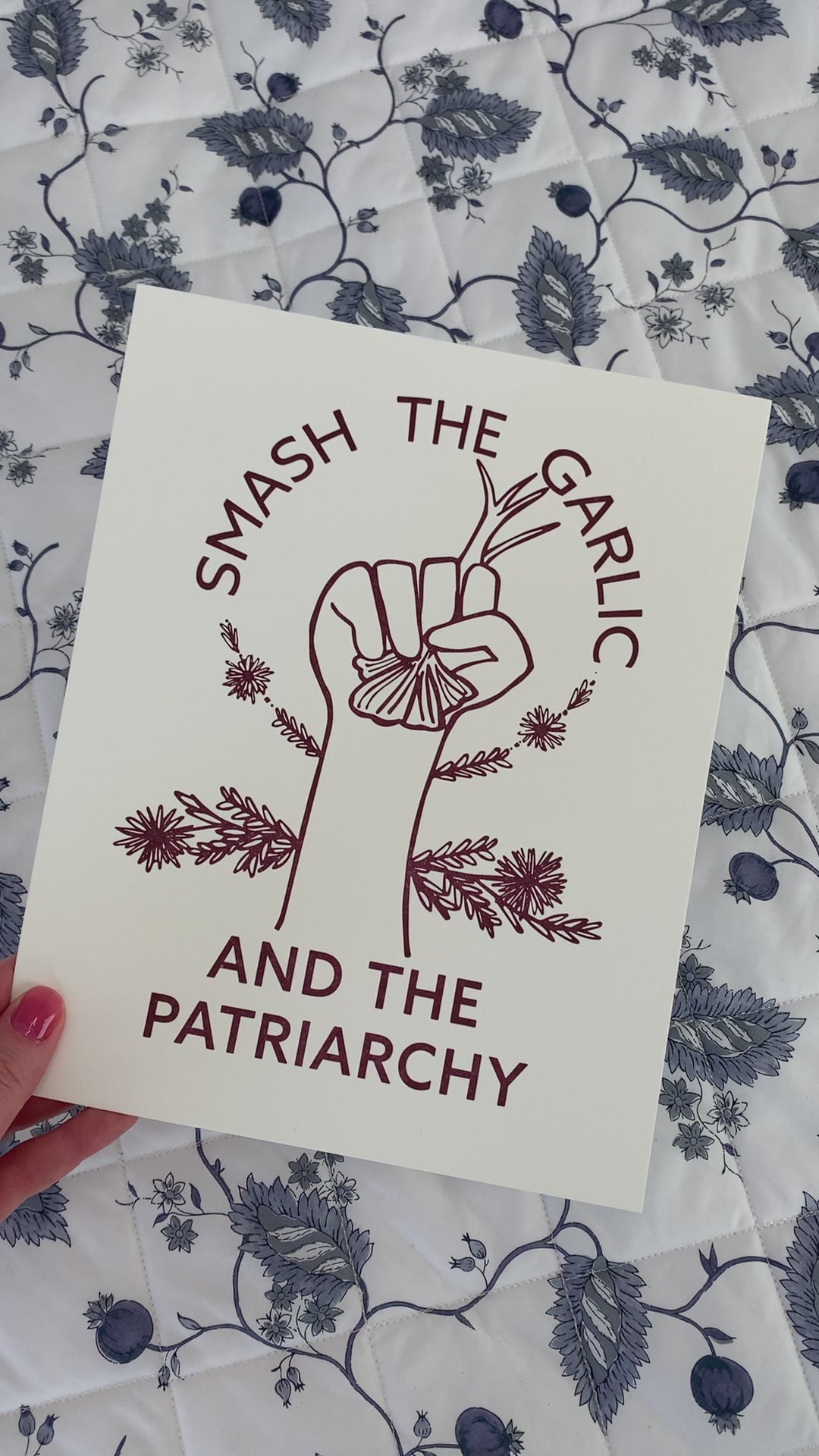 An art print that reads "Smash the Garlic and the Patriarchy" in a dark red color with an illustration of a hand holding garlic