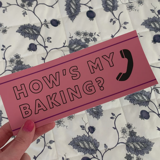 A woman holds a bumper sticker that reads "Hows my baking?"