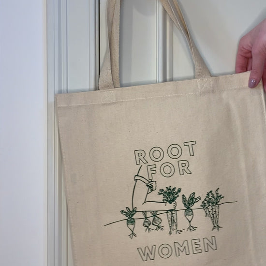A canvas tote with "Root for Women" in green block letters hangs on a doorknob