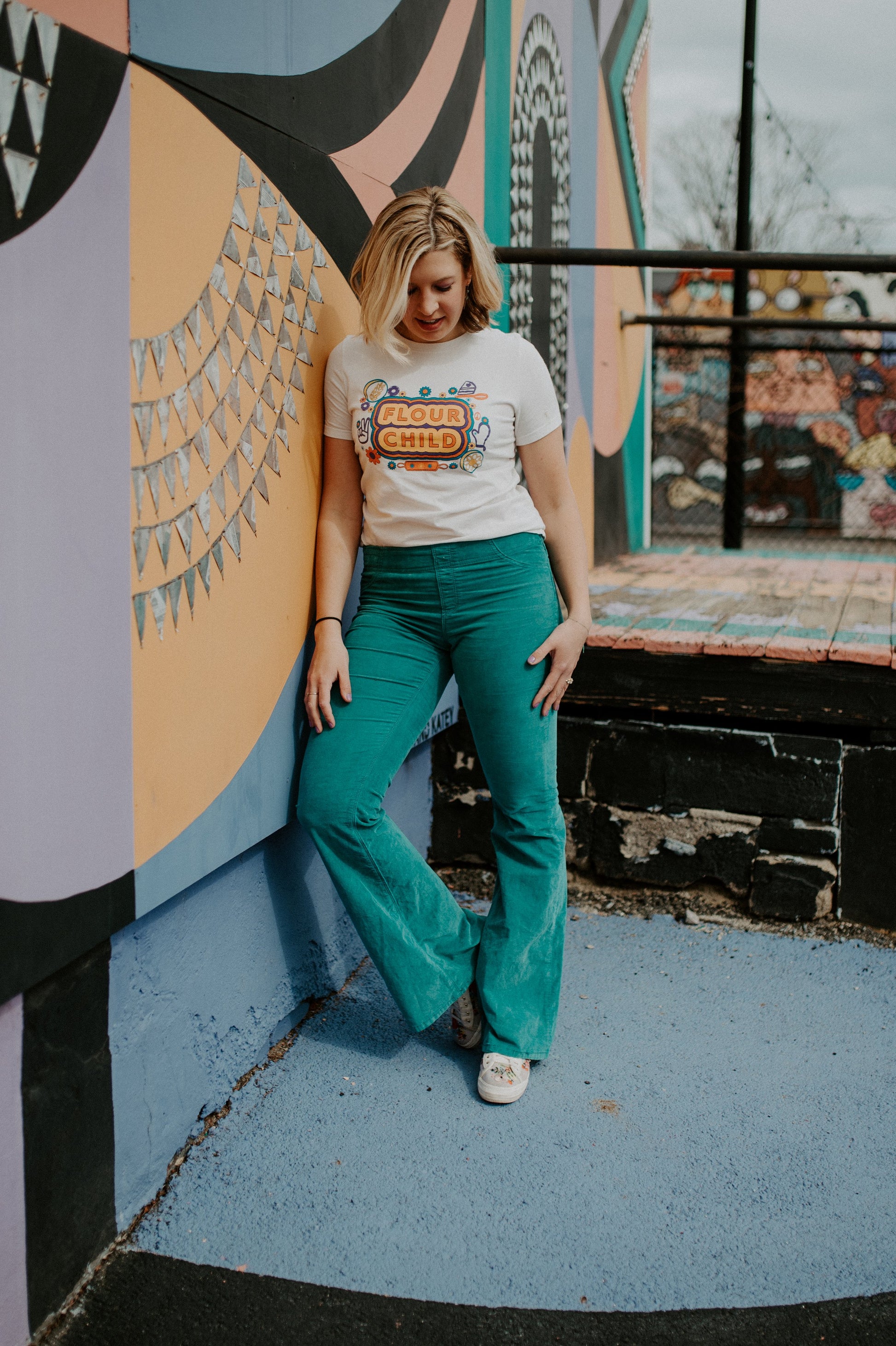 A woman in a white Flour Child tee and teal pants leans against a mural