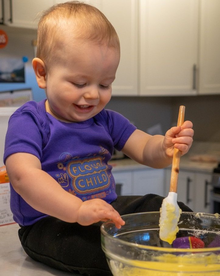 A baby wears the purple Flour Child onesie and holds a spatula over a bowl