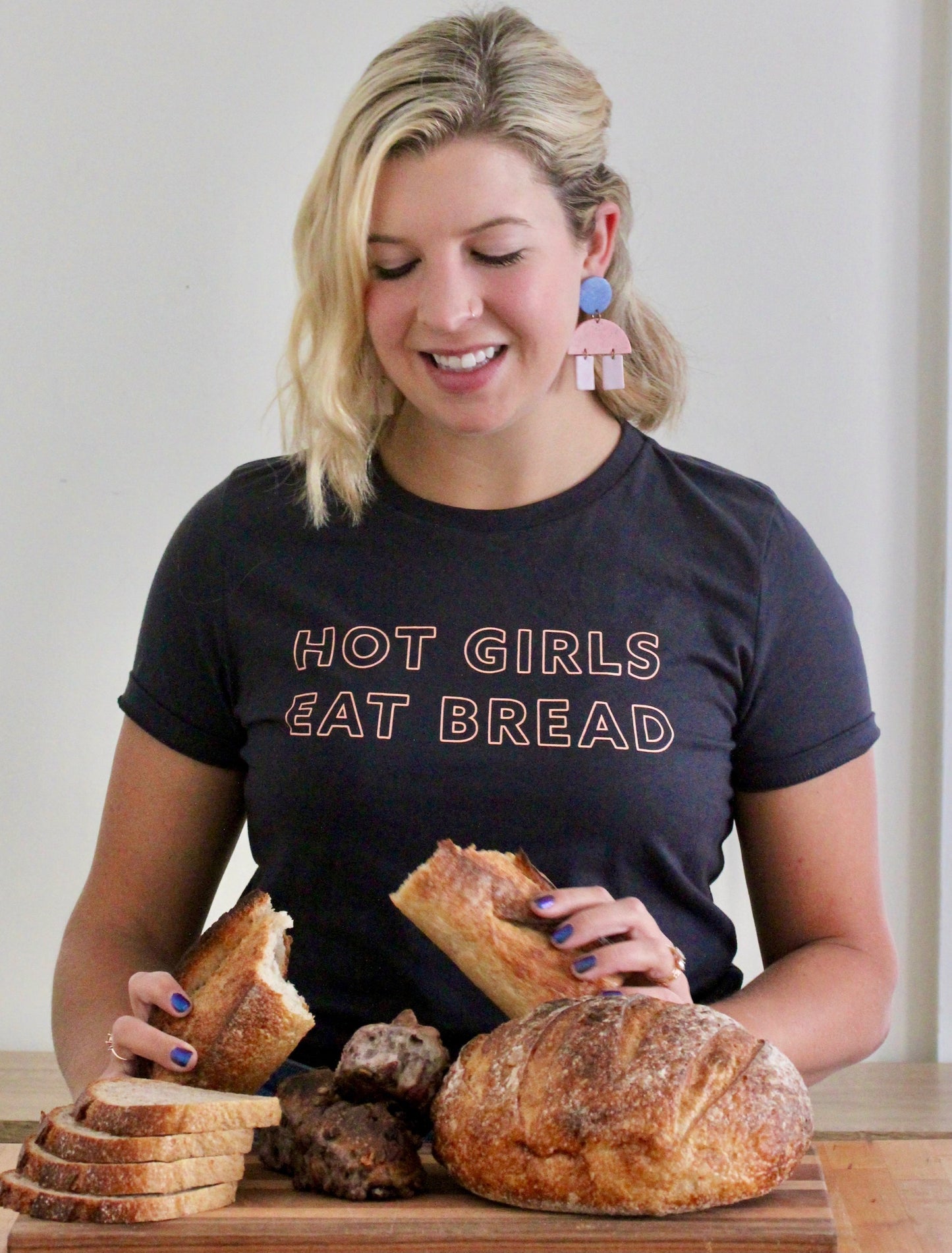 A woman in a dark grey tee that reads "Hot Girls Eat Bread" looks down at loaves and slices of bread
