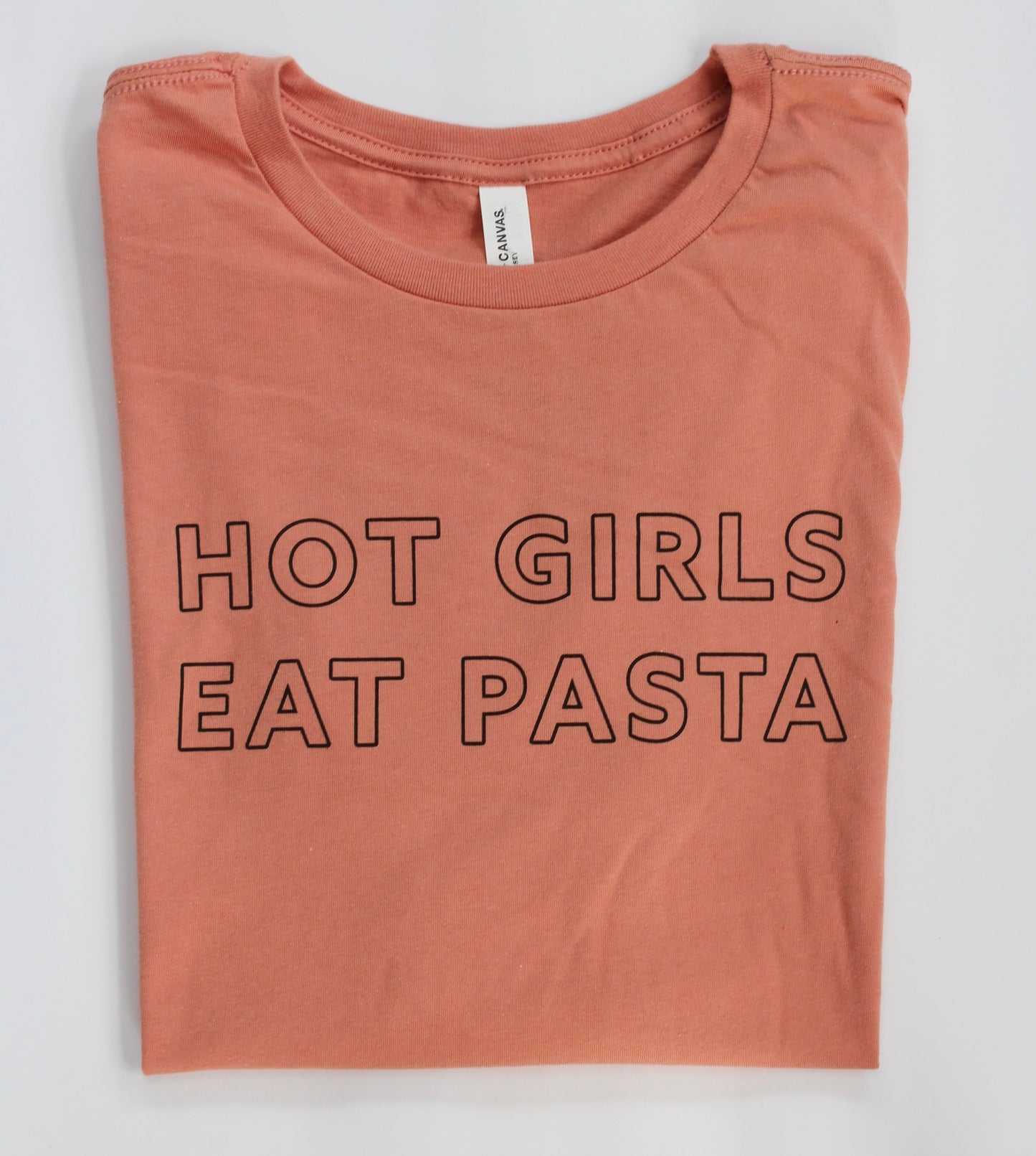 A folded peach tee with the words Hot Girls Eat Pasta in black block letters