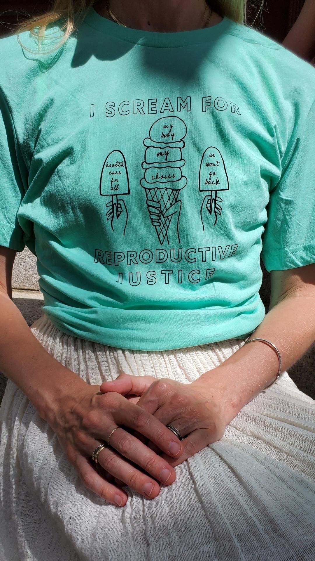 A woman wears a mint green tee that reads "I Scream for Reproductive Justice" with a white skirt