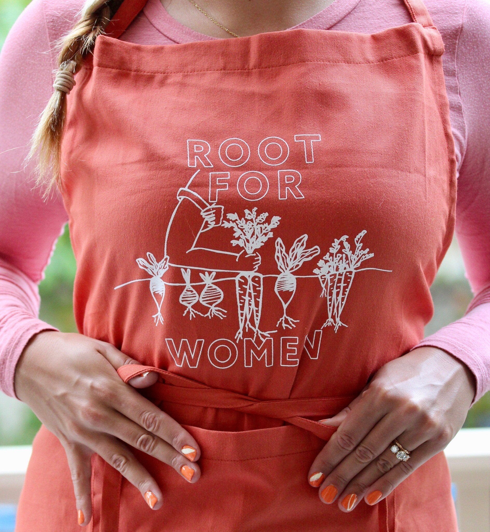 A woman wears a "Root for Women" apron over a pink top