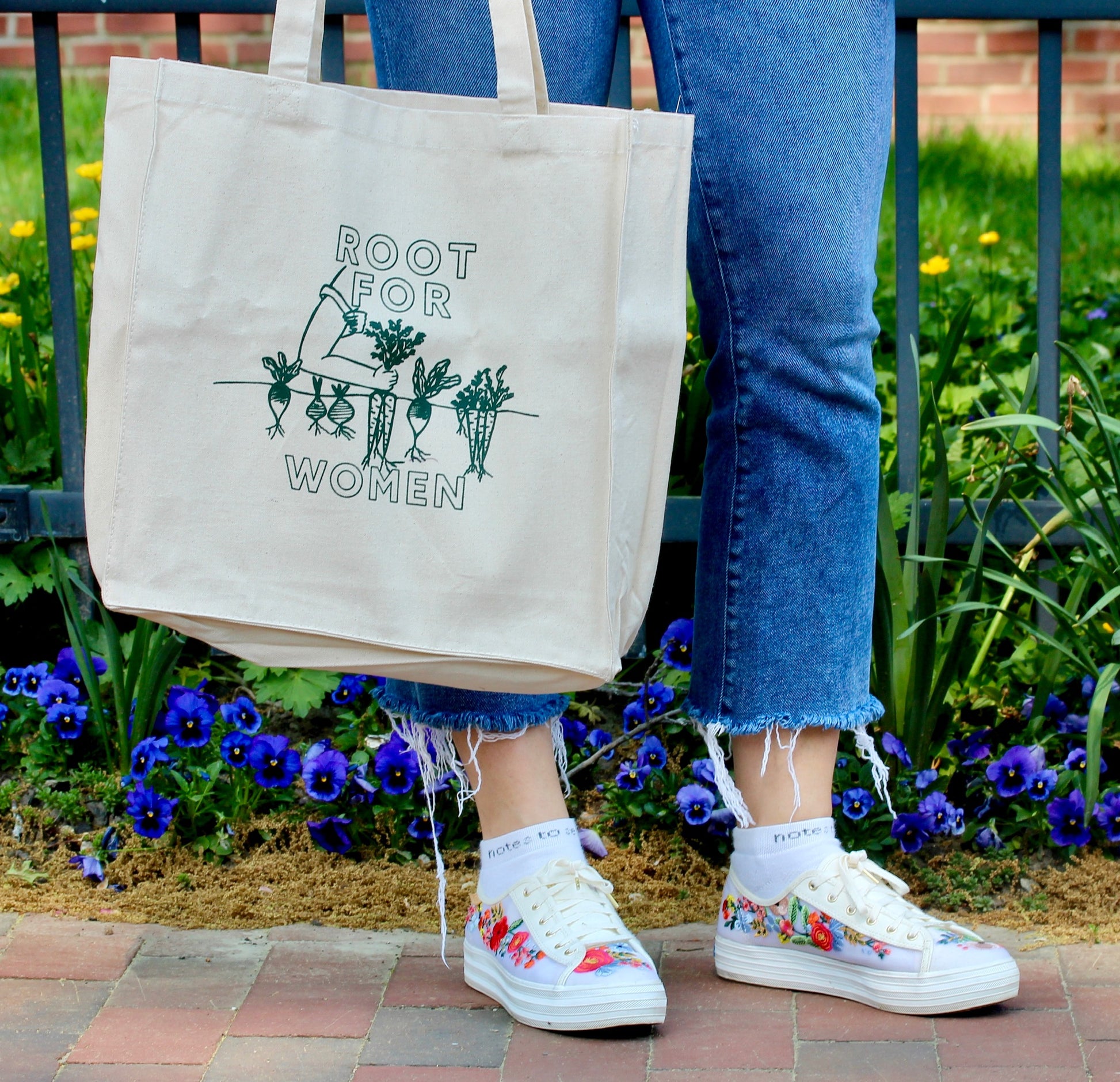 A woman holds a canvas tote that reads "Root for Women" in green block letters