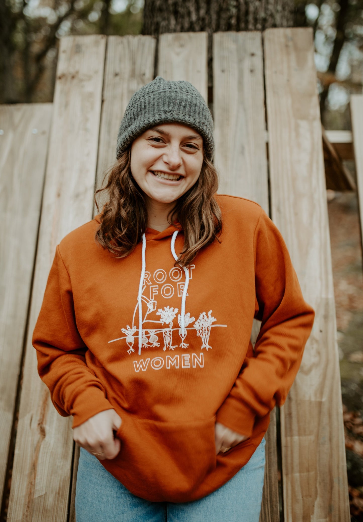 A woman wears a "Root for Women" hoodie with a green beanie and jeans