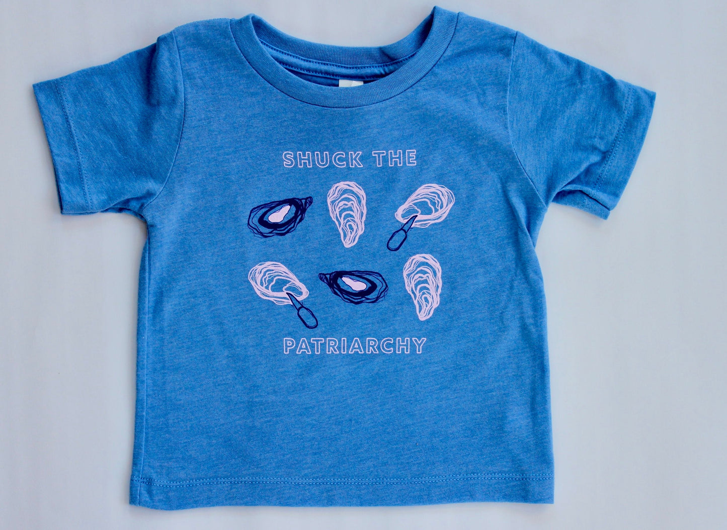 A baby sized tee in bright blue reads "Shuck the Patriarchy" in block letters with oyster illustrations