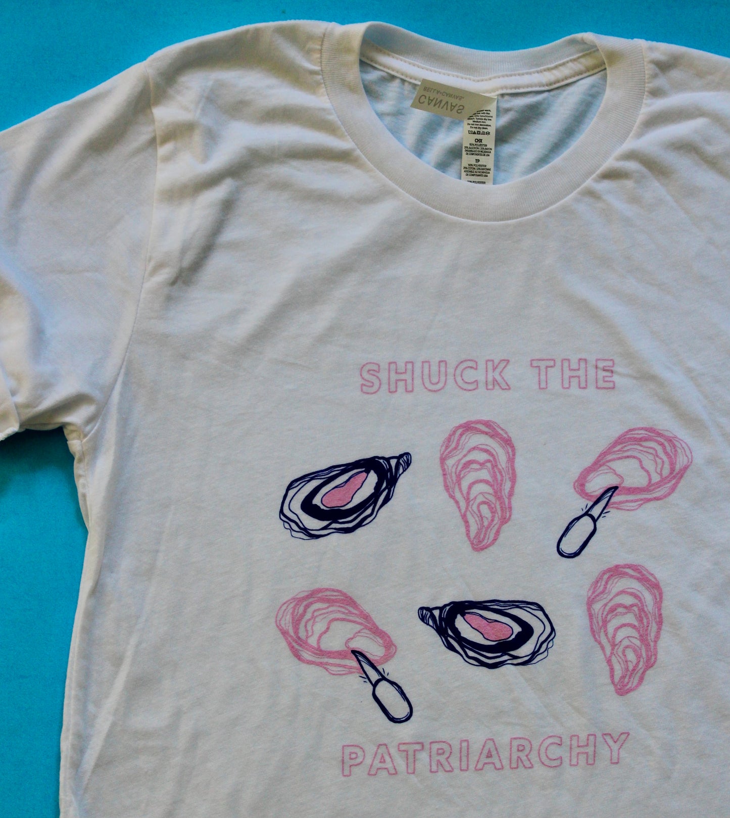 A white tee reads "Shuck the Patriarchy" in pink block letters with oyster illustrations