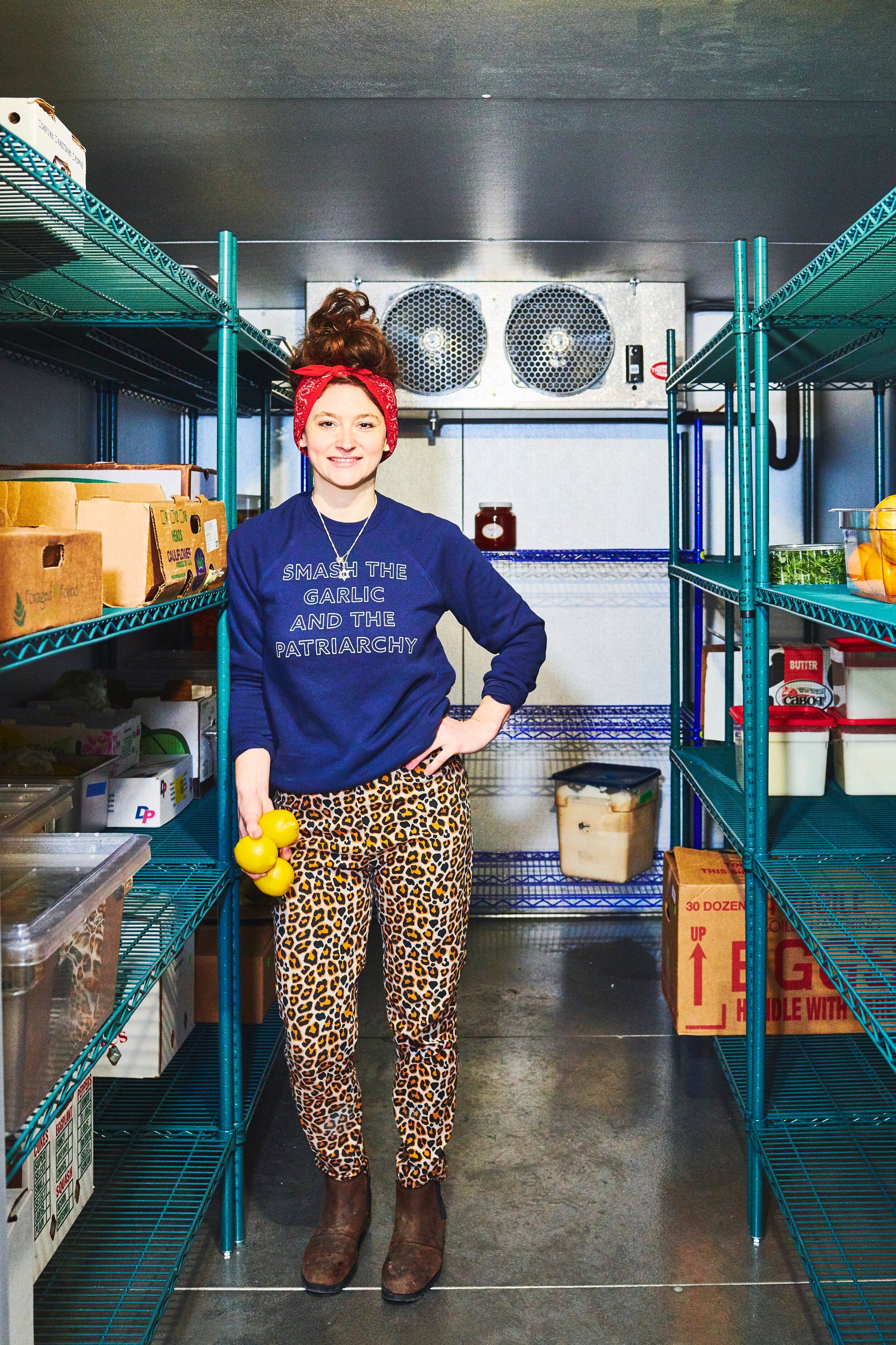 A woman wearing a navy blue "Smash the Garlic and the Patriarchy" stands in a walk-in refrigerator