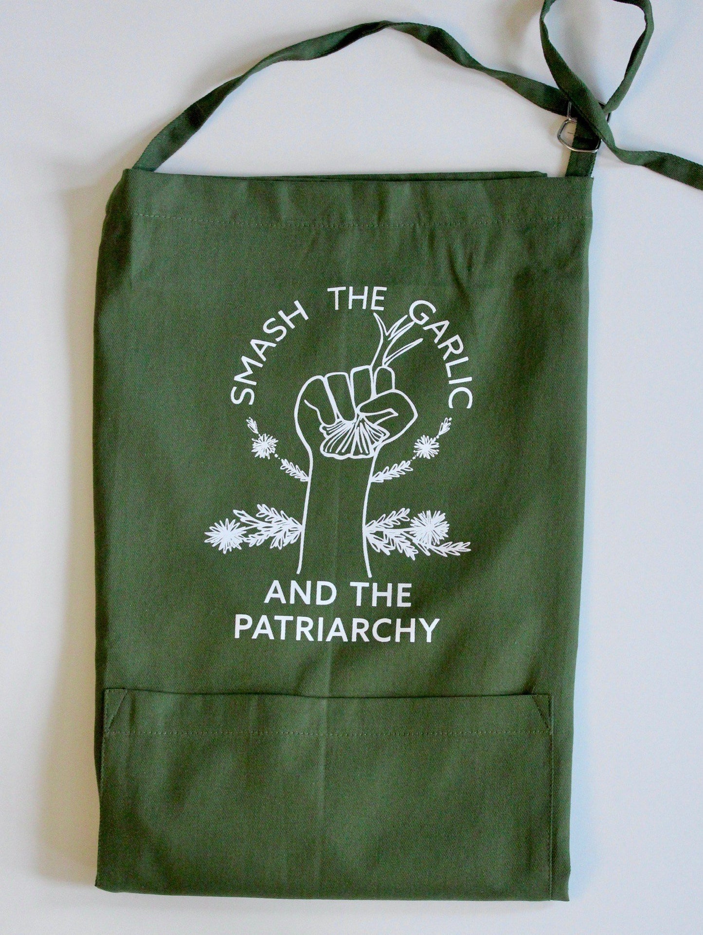 A folded dark green apron with the words "Smash the Garlic and the Patriarchy" and an illustration of a hand holding garlic