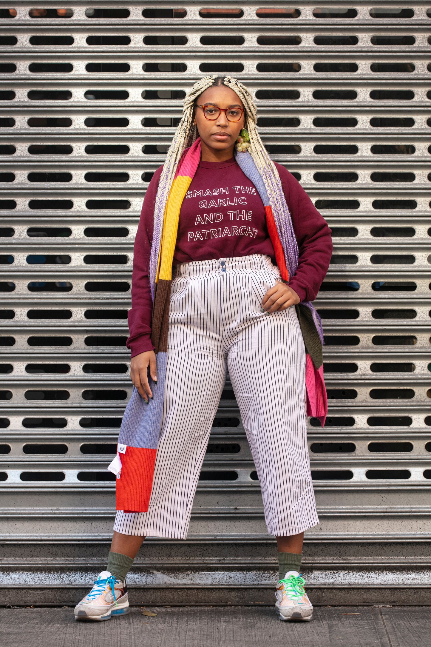 A woman wears a maroon "Smash the Garlic and the Patriarchy" crewneck with a colorful scarf and white striped pants