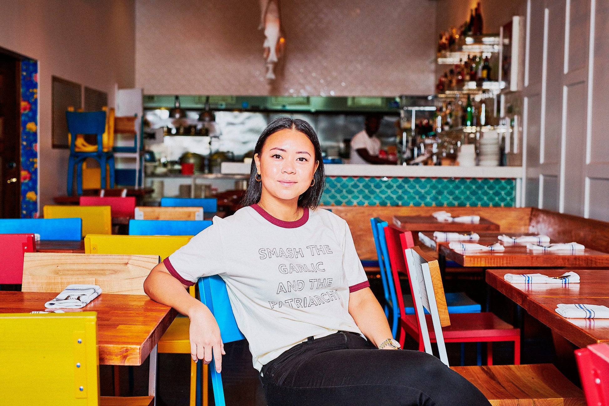 A woman wearing a "Smash the Garlic and the Patriarchy" and black jeans sits in a colorful restaurant