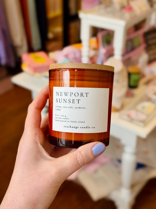 Hand holding a 'Newport Sunset' candle with a blurred indoor background