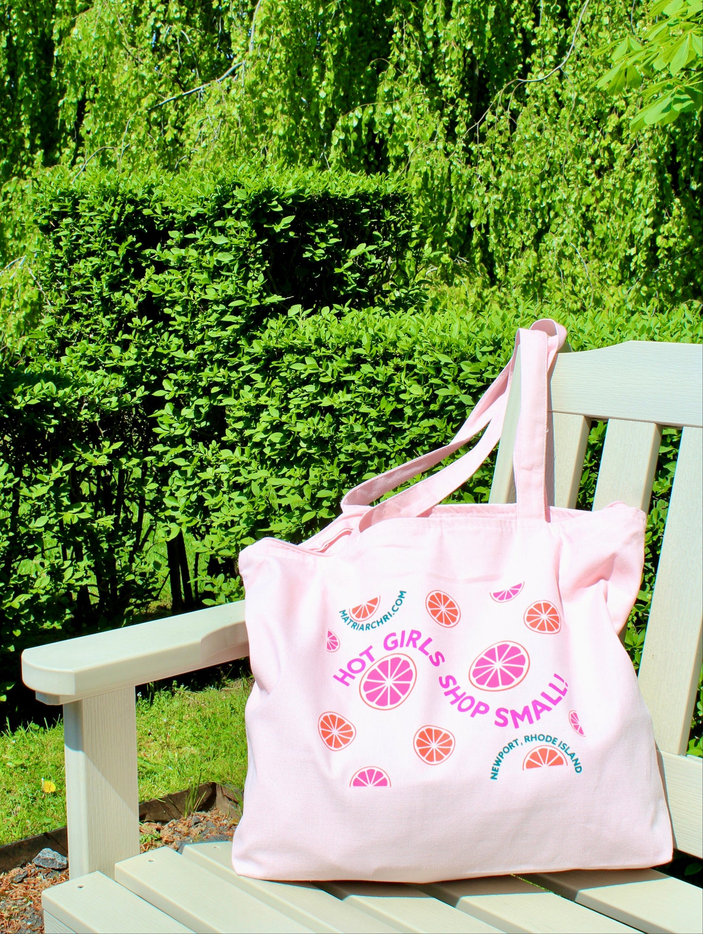Pink tote bag with citrus designs and text reading "Hot Girls Shop Small!" on a wooden bench in a garden setting
