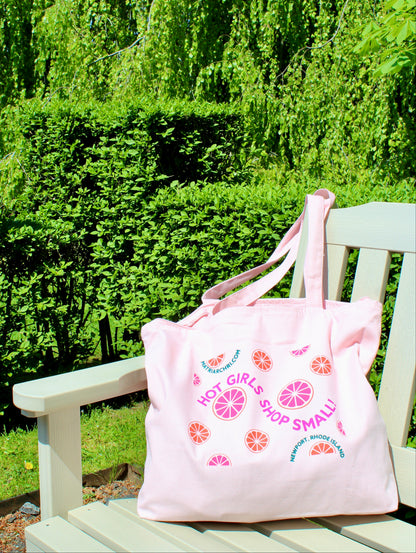 Pink tote bag with citrus designs and text reading "Hot Girls Shop Small!" on a wooden bench in a garden setting