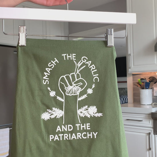 A green apron that reads "Smash the Garlic and the Patriarchy" in white letters hangs on a hanger