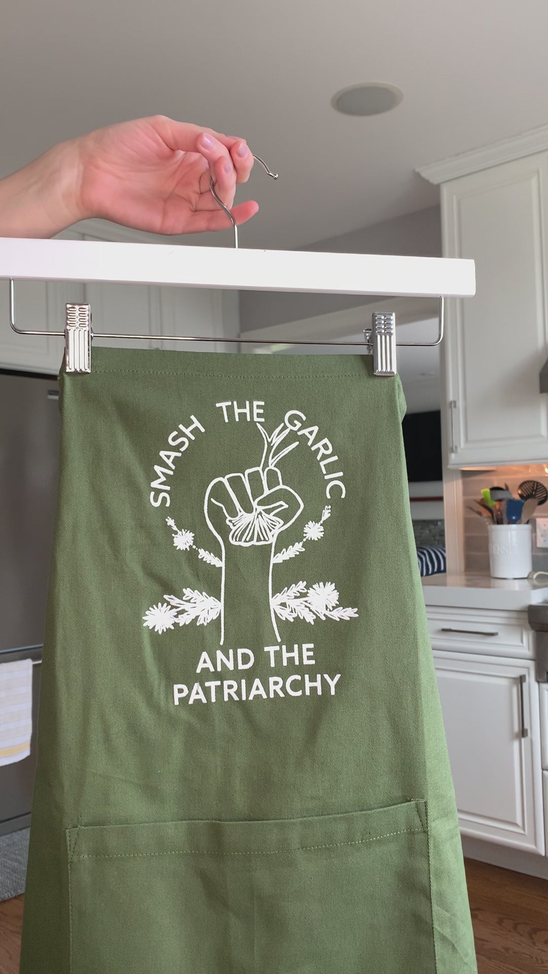 A green apron that reads "Smash the Garlic and the Patriarchy" in white letters hangs on a hanger