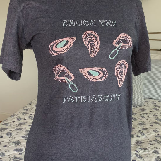 A heather navy tee that reads "Shuck the Patriarchy" hangs on a manikin