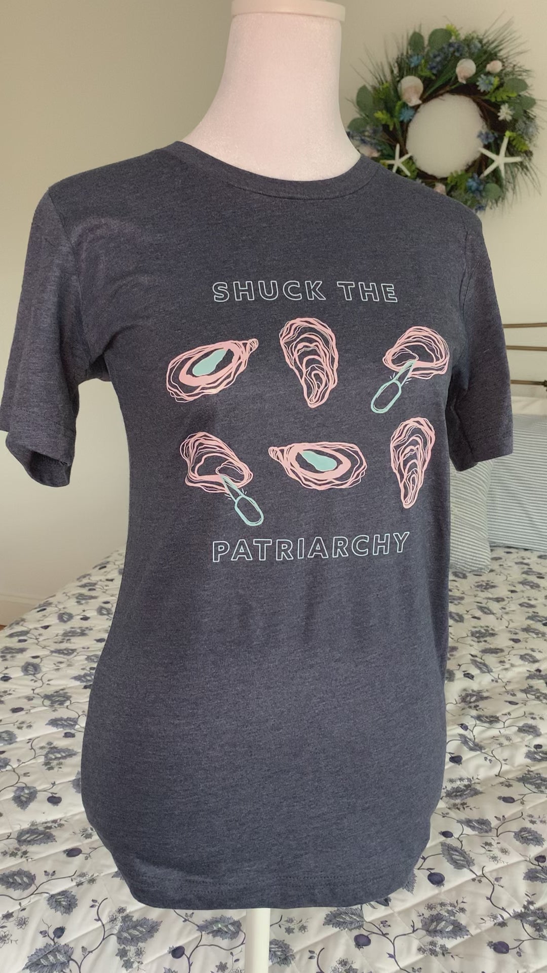 A heather navy tee that reads "Shuck the Patriarchy" hangs on a manikin