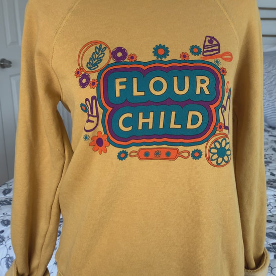 A yellow crewneck sweatshirt that reads "Flour Child" hangs on a manikin 