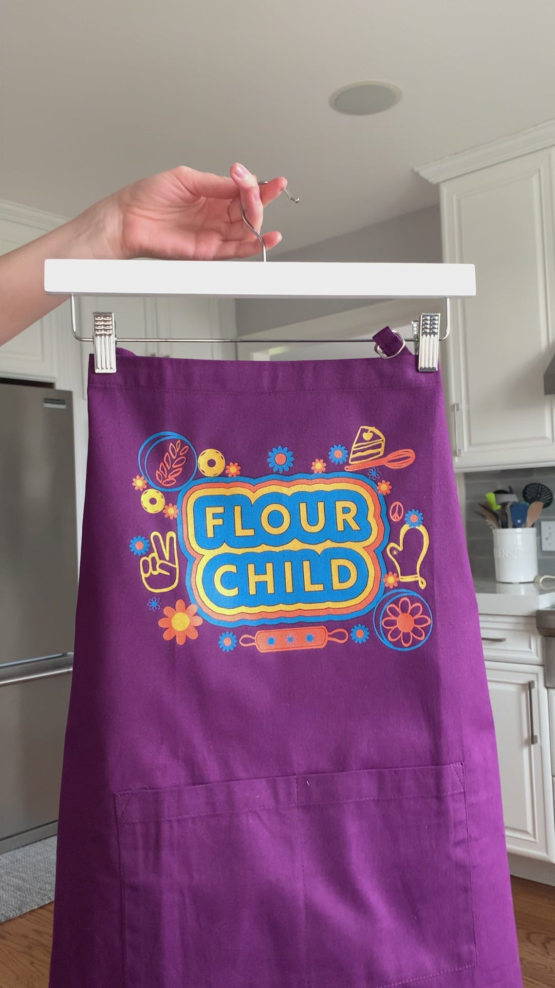 A purple apron that reads "Flour Child" with colorful illustrations hangs on a hanger
