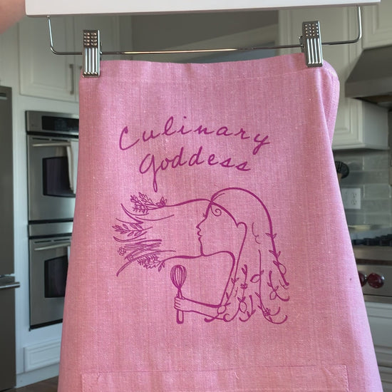 A pink chambray apron with the words "Culinary Goddess" hangs on a hanger