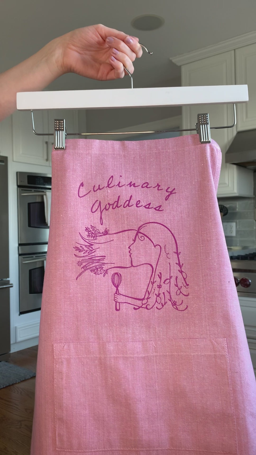 A pink chambray apron with the words "Culinary Goddess" hangs on a hanger