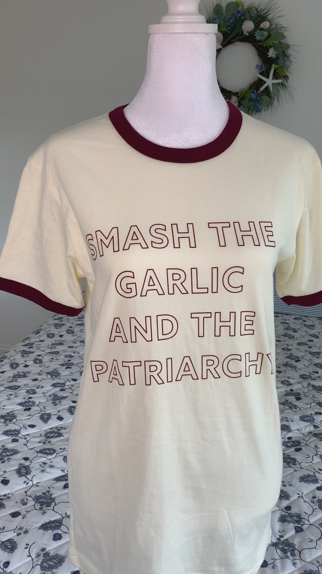 An off-white ringer tee that reads "Smash the Garlic and the Patriarchy" hangs on a manikin 