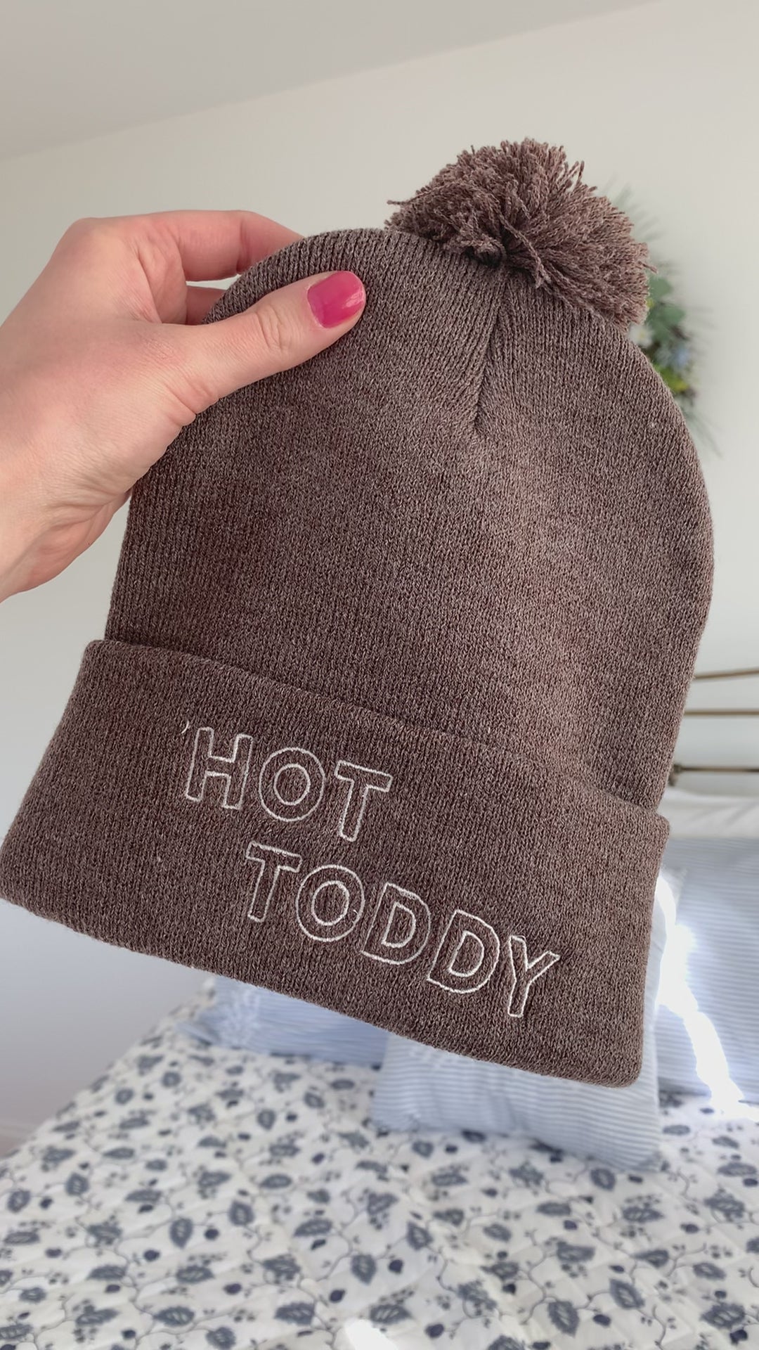 A woman holds a brown beanie with a pom pom that reads "Hot Toddy"