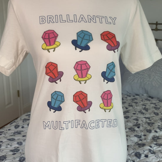 A white t-shirt that reads "Brilliantly Multifaceted" hangs on a manikin