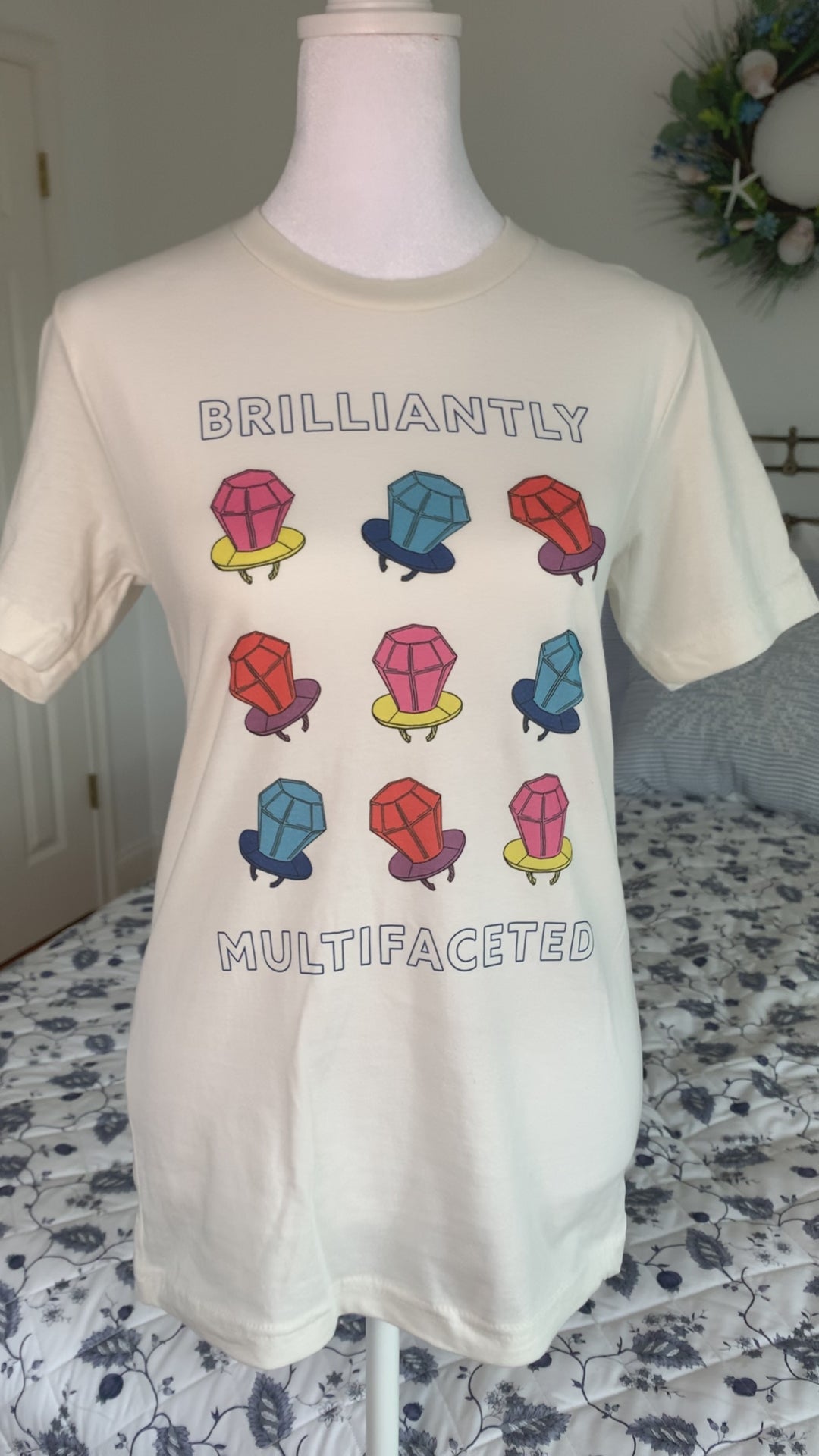A white t-shirt that reads "Brilliantly Multifaceted" hangs on a manikin