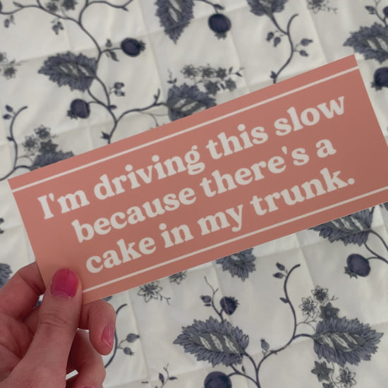 A woman holds a bumper sticker that reads "I'm driving this slow because there's a cake in my trunk."