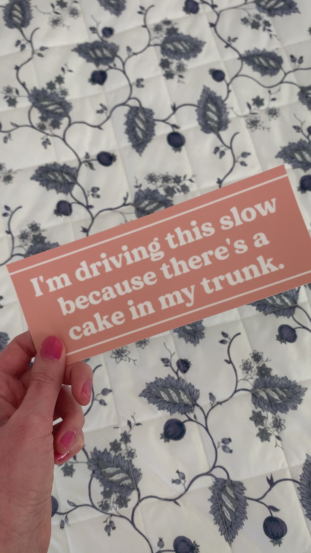 A woman holds a bumper sticker that reads "I'm driving this slow because there's a cake in my trunk."