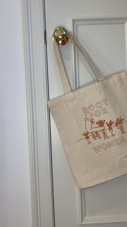 A canvas tote with the words "Root for Women" in orange block letters hangs on a doorknob