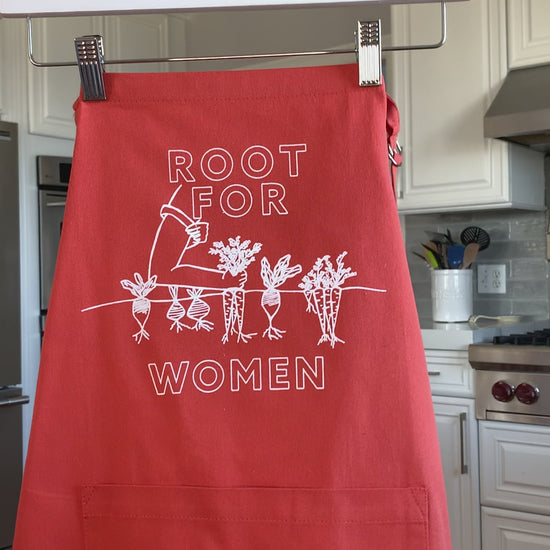 An orange apron that reads "Root for Women" in white block letters hangs on a hanger
