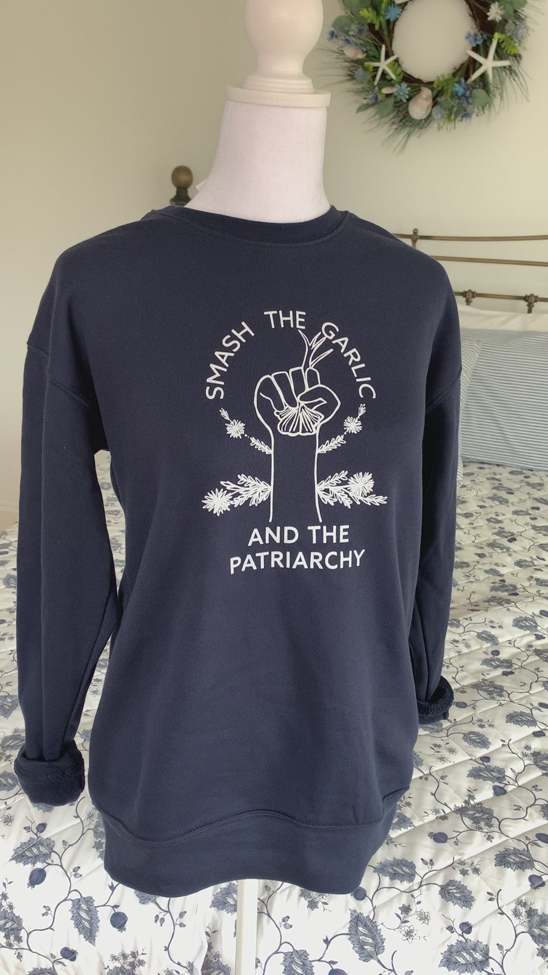 A navy crewneck that reads "Smash the Garlic and the Patriarchy" with a garlic illustration hangs on a manikin